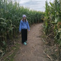 A woman in a hoodie and long skirt stands on a dirt pathway inside a tall corn maze. The corn stalks rise high on both sides, and the sky is overcast above.