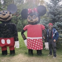 A person stands beside two large hay sculptures designed to resemble Mickey Mouse and Minnie Mouse. The figures are made from stacked hay bales with painted clothing and large black circular ears. They are displayed outdoors on a grassy area with evergreen trees in the background.