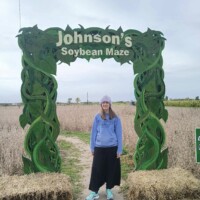 A person stands beneath a green decorative archway that reads “Johnson’s Soybean Maze.” The arch is designed with winding vine shapes. The person is wearing a light blue hoodie, black skirt, knit hat, and sneakers. Behind the arch is a dry soybean field under an overcast sky, with straw bales on the ground at the entrance.