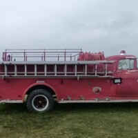 A vintage red fire truck is parked on a grassy field under an overcast sky. The truck has a long ladder on top and equipment along the sides. A stack of hay bales and a pumpkin display are visible in the distance behind it.