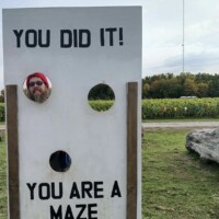 A person sticks their face through a circular cutout in a white photo board that reads “You did it! You are a maze master.” Cornfields and trees are visible in the background under an overcast sky.
