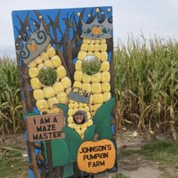 A person poses with their face in a cutout board at Johnson’s Pumpkin Farm. The board is painted to look like two corn characters wearing crowns, with a sign that says “I am a maze master” next to a corn maze in the background.