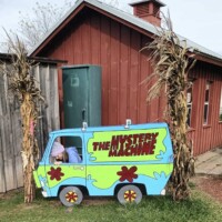 A person crouches behind a painted wooden cutout of the Mystery Machine van from Scooby-Doo, adjusting their position so their feet are hidden. The cutout is set up outdoors between two cornstalk decorations in front of a red wooden farm building.