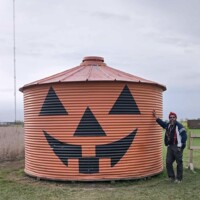 A person stands beside a round metal grain silo painted orange with a large black jack-o’-lantern face on the side. The person is wearing a red knit hat and a blue jacket. The silo sits on a grassy field under an overcast sky.