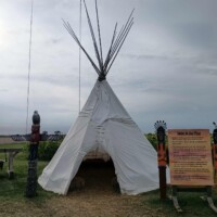 A white canvas tipi-style structure stands on a grassy area under an overcast sky. The entrance is open, showing hay inside. A tall wooden carved figure is positioned on the left, and an informational sign stands on the right. Additional carved wooden decorations are visible behind the sign.