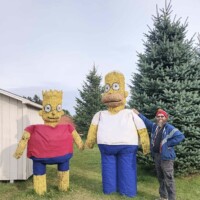 A person stands next to two large hay sculptures designed to look like Bart and Homer Simpson at Johnson’s Pumpkin Farm. The figures are made from stacked hay bales with painted clothing and are positioned on a grassy area near evergreen trees.