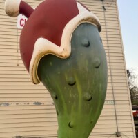 Close-up of the World’s Largest Christmas Pickle statue in a Santa hat at Christmas Winter Wonderland in Fort Smith, Arkansas, photographed outdoors before dark with the building siding behind it.