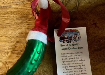 Green pickle Christmas ornament wearing a Santa hat with a tag explaining the Christmas pickle tradition, sitting on a wooden surface.