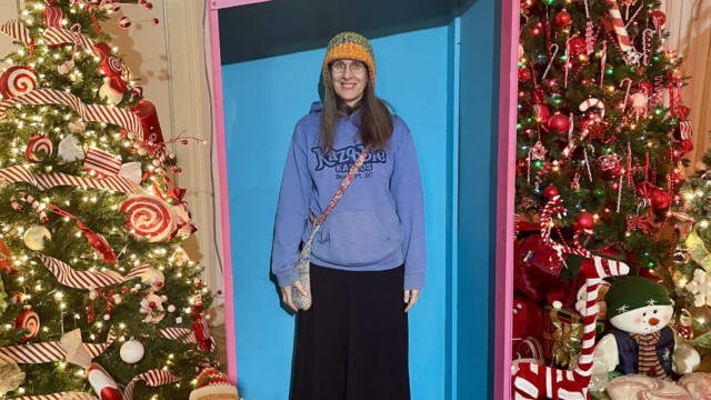Woman standing inside a life-sized Barbie doll box display at Christmas Winter Wonderland in Fort Smith, Arkansas, surrounded by decorated Christmas trees and candy-themed holiday décor.