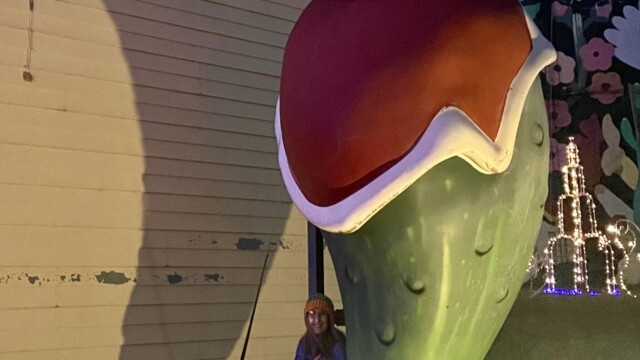 Woman standing beneath a giant Christmas pickle statue wearing a Santa hat at night in Fort Smith, Arkansas, with holiday lights and decorations glowing around it.