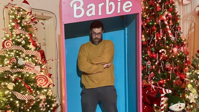 Man standing inside a life-sized Barbie doll box display at Christmas Winter Wonderland in Fort Smith, Arkansas, surrounded by decorated Christmas trees and candy-themed holiday décor.