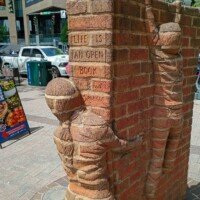 A close-up of the "Life is an Open Book" brick sculpture by Brad Spencer, located in uptown Charlotte, NC. Two child figures sculpted from brick climb the spine of a massive open book. The words "Life is an Open Book" are carved into the bricks, with the artist's signature "Brad Spencer '02" etched below. A busy city sidewalk, street signs, and downtown buildings are visible in the background.