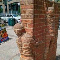 A close-up of the "Life is an Open Book" brick sculpture by Brad Spencer, located in uptown Charlotte, NC. Two child figures sculpted from brick climb the spine of a massive open book. The words "Life is an Open Book" are carved into the bricks, with the artist's signature "Brad Spencer '02" etched below. A busy city sidewalk, street signs, and downtown buildings are visible in the background.