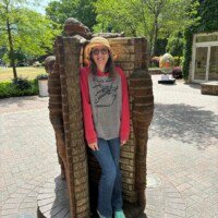 A woman (Nancy) stands tucked inside the open pages of the "Life is an Open Book" brick sculpture by Brad Spencer in uptown Charlotte, NC. The sculpture's brick pages frame her on both sides, with brick-carved child figures climbing above her. She is smiling, wearing a straw hat, sunglasses, a Star Wars hoodie, jeans, and teal sneakers. A courtyard with trees and a building entrance is visible in the background.