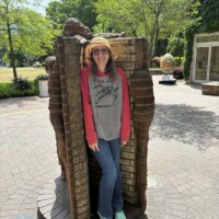 A woman (Nancy) stands tucked inside the open pages of the "Life is an Open Book" brick sculpture by Brad Spencer in uptown Charlotte, NC. The sculpture's brick pages frame her on both sides, with brick-carved child figures climbing above her. She is smiling, wearing a straw hat, sunglasses, a Star Wars hoodie, jeans, and teal sneakers. A courtyard with trees and a building entrance is visible in the background.