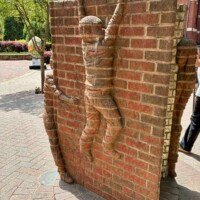 A side view of the "Life is an Open Book" brick sculpture by Brad Spencer in uptown Charlotte, NC. A brick-carved child figure is shown mid-climb on the spine of the open book, arms reaching upward and legs braced against the surface. A second smaller figure is partially visible at the bottom left. The Ratcliffe's Flowers sign is visible in the background alongside street trees and a sidewalk.