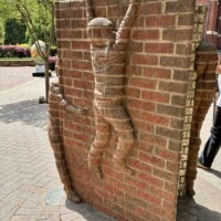 A side view of the "Life is an Open Book" brick sculpture by Brad Spencer in uptown Charlotte, NC. A brick-carved child figure is shown mid-climb on the spine of the open book, arms reaching upward and legs braced against the surface. A second smaller figure is partially visible at the bottom left. The Ratcliffe's Flowers sign is visible in the background alongside street trees and a sidewalk.