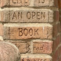 A close-up of the brick spine of the "Life is an Open Book" sculpture in uptown Charlotte, NC, showing the title and artist signature carved directly into the bricks. The words "Life is an Open Book" are etched across three rows, with "Brad Spencer '02" carved below. The rounded brick head of one of the child figures is partially visible at the lower left.