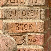 A close-up of the brick spine of the "Life is an Open Book" sculpture in uptown Charlotte, NC, showing the title and artist signature carved directly into the bricks. The words "Life is an Open Book" are etched across three rows, with "Brad Spencer '02" carved below. The rounded brick head of one of the child figures is partially visible at the lower left.
