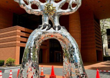 A full view of the Firebird sculpture outside the Bechtler Museum of Modern Art at 420 South Tryon Street in Charlotte, NC. The large abstract sculpture is covered entirely in mosaic mirror tiles that reflect light in all directions. It stands on two arched legs forming a tunnel beneath its body, with outstretched wings and a crown of gold mosaic spikes at the top. A gold sunburst medallion decorates the chest. The sculpture is surrounded by orange traffic cones and rope barriers due to damage at the base. The warm brick facade of the Bechtler Museum is visible in the background.