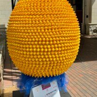 A large decorative egg sculpture covered entirely in small yellow rubber ducks, part of the Easter Eggs on Parade public art installation in uptown Charlotte, NC. The egg is mounted on a stand with a ruff of blue tulle at its base. Hundreds of tiny rubber ducks cover every inch of the surface, all facing outward. A white informational placard is visible at the base identifying it as part of the Charlotte SHOUT! Festival. A brick building entrance is visible in the background.