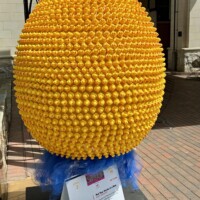 A large decorative egg sculpture covered entirely in small yellow rubber ducks, part of the Easter Eggs on Parade public art installation in uptown Charlotte, NC. The egg is mounted on a stand with a ruff of blue tulle at its base. Hundreds of tiny rubber ducks cover every inch of the surface, all facing outward. A white informational placard is visible at the base identifying it as part of the Charlotte SHOUT! Festival. A brick building entrance is visible in the background.