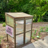 A small wooden and clear panel community sharing box in a residential yard in Charlotte, NC, resembling a miniature greenhouse structure. A pink handwritten sign on the front reads "Free Goods" with smaller text reading "Fresh from our Garden." The box contains several labeled bins visible through the clear panels. It is surrounded by a garden bed with mulch, green plants, and emerging foliage.