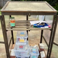 The interior of a community free goods sharing box in Charlotte, NC, with its doors open to reveal the contents. The upper shelf holds a labeled jar of dog treats, a measuring cup, fabric tote bags, and loose papers. The lower shelf contains several clear plastic bins with handwritten colored labels, including categories for stationery and crafts, toys, books, and seeds, all stacked and organized on brick risers.