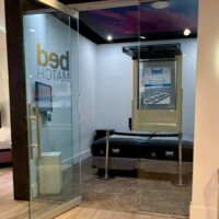 Glass-enclosed BedMatch room at Furnitureland South, with the sensor bed visible inside and the brand logo etched on the glass door