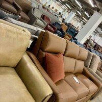 Tan leather sofas with a rust-colored accent pillow displayed in the Furnitureland South outlet, with rows of additional furniture stretching into the background