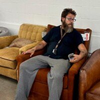 A man with a beard sits in a brown leather recliner inside the Furnitureland South outlet, wearing a navy polo and lanyard, with a tan chair beside him