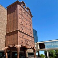 Full view of the World's Largest Highboy Dresser at Furnitureland South, towering several stories tall with carved drawers and ball-and-claw feet, set against the building and glass skybridge