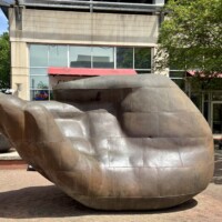 A wide shot of a large bronze hand sculpture on a city sidewalk in uptown Charlotte, NC. The hand is oriented palm-up with fingers slightly curled, rendered in smooth bronze panels with visible seam lines across the surface. The sculpture is large enough for a person to sit inside the palm. Little Village Grill restaurant with its red awning is visible in the background, along with downtown Charlotte buildings and street trees.