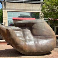 A wide shot of a large bronze hand sculpture on a city sidewalk in uptown Charlotte, NC. The hand is oriented palm-up with fingers slightly curled, rendered in smooth bronze panels with visible seam lines across the surface. The sculpture is large enough for a person to sit inside the palm. Little Village Grill restaurant with its red awning is visible in the background, along with downtown Charlotte buildings and street trees.