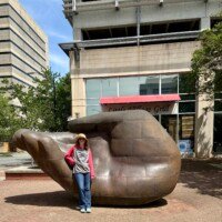 A woman (Nancy) stands in front of the large bronze hand sculpture on a city sidewalk in uptown Charlotte, NC, showing the scale of the piece — the hand reaches well above her head. She is wearing a straw hat, sunglasses, a Star Wars hoodie, jeans, and teal sneakers. Little Village Grill with its red awning is visible behind her, along with downtown Charlotte buildings and trees.