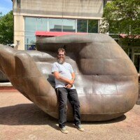 A man (Zach) stands in front of the large bronze hand sculpture on a city sidewalk in uptown Charlotte, NC, smiling with his hands clasped in front of him. The sculpture towers behind him, its palm-up fingers visible over his shoulder. He is wearing a white t-shirt, black pants, and green sneakers. Downtown Charlotte buildings and trees are visible in the background.