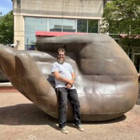 A man (Zach) stands in front of the large bronze hand sculpture on a city sidewalk in uptown Charlotte, NC, smiling with his hands clasped in front of him. The sculpture towers behind him, its palm-up fingers visible over his shoulder. He is wearing a white t-shirt, black pants, and green sneakers. Downtown Charlotte buildings and trees are visible in the background.