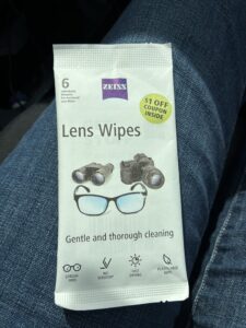 A small sample packet of ZEISS Lens Wipes resting on a person's lap in a vehicle, with denim jeans visible beneath it. The white packet contains six individually wrapped pre-moistened lens wipes and features a $1 off coupon badge in the upper right corner. The packaging shows illustrations of binoculars, a camera, and eyeglasses, with the tagline "Gentle and thorough cleaning" and icons for streak-free, no scratch, fast drying, and plastic-free features.