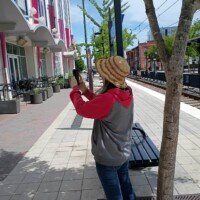 A woman (Nancy) photographs the exterior of Seventh Street Station in uptown Charlotte, NC, holding her phone up toward the building. She is wearing a straw hat, a Star Wars hoodie, and jeans, standing on the sidewalk beside a young street tree and a black bench. The light rail tracks, additional street trees, and colorful building facades are visible stretching down the block behind her.
