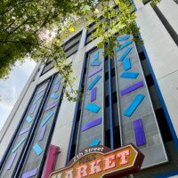 An upward-looking view of the exterior of Seventh Street Station in uptown Charlotte, NC, home to the Musical Parking Garage. The building facade features bold blue trim and decorative blue and purple geometric tile panels arranged in vertical columns across multiple floors. The 7th Street Public Market sign is visible at the bottom of the frame in orange and red lettering. Tree branches with green leaves frame the upper portion of the image against a partly cloudy sky.