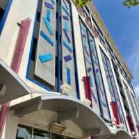 An upward-angled view of the front entrance facade of Seventh Street Station in uptown Charlotte, NC. The building features tall vertical columns of blue and teal geometric tile panels set against a white exterior with blue trim and deep red accent panels. A curved white awning extends over the ground floor entrance below. Tree branches are visible at the upper right against a bright blue sky.