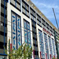 A straight-on view of the full exterior of Seventh Street Station, the Musical Parking Garage in uptown Charlotte, NC. The multi-story building features a white facade with bold blue trim and columns of blue geometric tile panels running vertically between the parking levels. The red "Seventh Street Station" sign is visible on the upper right of the building. Street trees and a light pole are in the foreground against a bright blue sky.