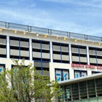 A wide street-level view of Seventh Street Station in uptown Charlotte, NC, showing the full length of the building from across the street. The multi-story parking structure features a white exterior with blue trim and the red "Seventh Street Station" sign spanning the upper right facade. A row of yellow dome-shaped light fixtures lines the roofline. The adjacent light rail station canopy is visible at the lower right, with mature trees lining the street in the foreground.