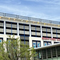 A wide street-level view of Seventh Street Station in uptown Charlotte, NC, showing the full length of the building from across the street. The multi-story parking structure features a white exterior with blue trim and the red "Seventh Street Station" sign spanning the upper right facade. A row of yellow dome-shaped light fixtures lines the roofline. The adjacent light rail station canopy is visible at the lower right, with mature trees lining the street in the foreground.