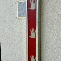 A close-up of one of the interactive hand panels on the exterior of the Musical Parking Garage at Seventh Street Station in uptown Charlotte, NC. The panel is a narrow vertical rectangle with a red background and three white hand silhouettes at different heights, designed to be touched to trigger musical notes. A small speaker is mounted on the concrete wall directly beside the panel. The surrounding wall is plain light gray concrete.