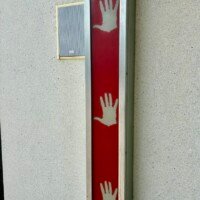 A close-up of one of the interactive hand panels on the exterior of the Musical Parking Garage at Seventh Street Station in uptown Charlotte, NC. The panel is a narrow vertical rectangle with a red background and three white hand silhouettes at different heights, designed to be touched to trigger musical notes. A small speaker is mounted on the concrete wall directly beside the panel. The surrounding wall is plain light gray concrete.