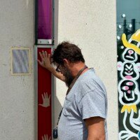 A man (Zach) presses his hand against one of the interactive musical panels on the exterior of the Musical Parking Garage at Seventh Street Station in uptown Charlotte, NC. He is leaning in slightly, wearing a white t-shirt, with visible tattoos on his forearm. The red panel with white hand silhouettes and the adjacent speaker are visible beside him. A colorful mural is partially visible on the wall to the right.
