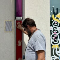 A man (Zach) presses his hand against one of the interactive musical panels on the exterior of the Musical Parking Garage at Seventh Street Station in uptown Charlotte, NC. He is leaning in slightly, wearing a white t-shirt, with visible tattoos on his forearm. The red panel with white hand silhouettes and the adjacent speaker are visible beside him. A colorful mural is partially visible on the wall to the right.