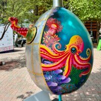 A large decorative egg sculpture painted with a vibrant underwater ocean scene, part of the Easter Eggs on Parade public art installation in uptown Charlotte, NC. The egg features a boldly colored pink and orange octopus with swirling tentacles surrounded by coral, sea anemones, and marine plants in shades of teal, purple, and green. A silver metal cap tops the egg, and a red octopus tentacle detail extends from the upper left side. The egg is mounted on a thin pole stand. Trees and a street scene are visible in the background.
