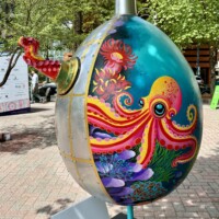 A large decorative egg sculpture painted with a vibrant underwater ocean scene, part of the Easter Eggs on Parade public art installation in uptown Charlotte, NC. The egg features a boldly colored pink and orange octopus with swirling tentacles surrounded by coral, sea anemones, and marine plants in shades of teal, purple, and green. A silver metal cap tops the egg, and a red octopus tentacle detail extends from the upper left side. The egg is mounted on a thin pole stand. Trees and a street scene are visible in the background.